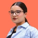 Portrait of Lydia Amazouz, a young woman with dark hair tied back, wearing glasses and a striped blue and white shirt, against a solid coral background.