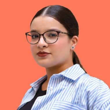 Portrait of Lydia Amazouz, a young woman with dark hair tied back, wearing glasses and a striped blue and white shirt, against a solid coral background.